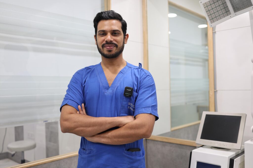 Dr. Shivam, dermatologist, standing in clinic wearing blue scrubs with advanced skin treatment equipment in background